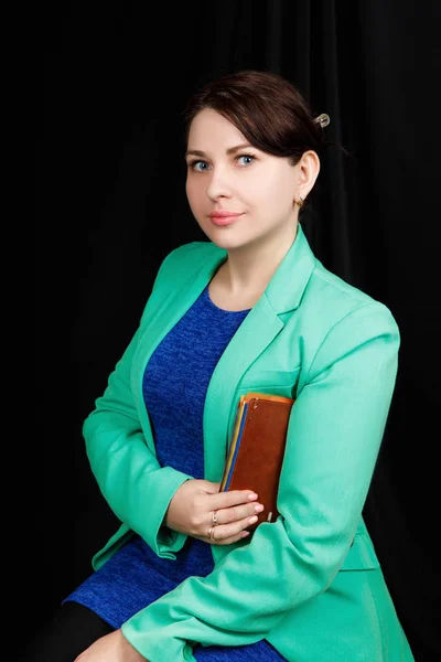 Studio  close up portrait of a girl of Slavic appearance worn a blue blouse and a blue-green jacket on a black background with brown notebook in her hands — 스톡 사진