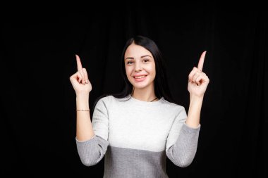 The concept of advertising. Positive, pretty, caucasion woman with a beaming smile and bright sweater on a black background indicates two index fingers and looks at the empty copy space 