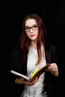 Slav görünümlü bir kızın yakın plan portresi elinde kahverengi defter olan siyah bir fonda siyah ceket giyiyor.