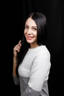 Portrait of serious beautiful female with black hair, has minimal makeup, looks calmly at camera, wears white jumper, stands against black background, being deep in thoughts