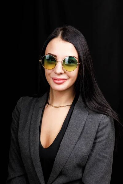 Image of sweet confident young lady standing over black background in studio, wearing white casual jacket and fashionable round green eyeglasses, having long dark hair. Youth concept — 스톡 사진