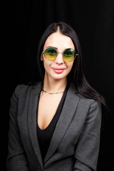 Image of sweet confident young lady standing over black background in studio, wearing white casual jacket and fashionable round green eyeglasses, having long dark hair. Youth concept — 스톡 사진