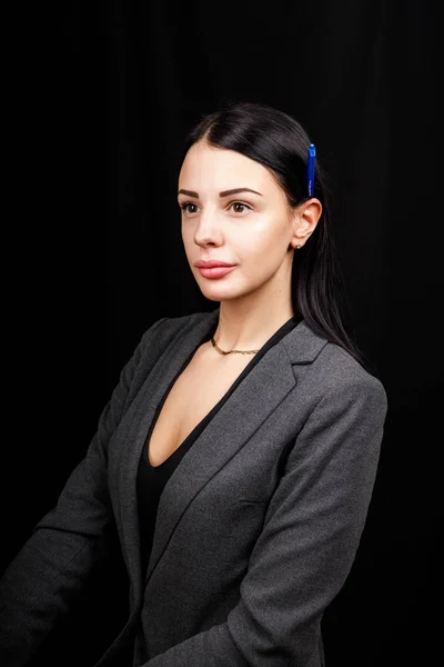 Retrato de jovem mulher de negócios em uma jaqueta cinza no fundo preto com caneta atrás da orelha — Fotografia de Stock