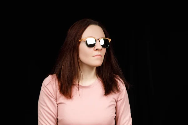 Bild einer süßen selbstbewussten jungen Frau, die vor schwarzem Hintergrund im Studio steht, rosa Bluse und modische Brille trägt und lange braune Haare hat — Stockfoto