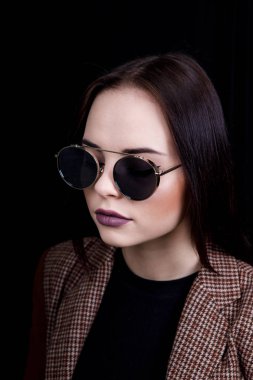 Stüdyoda siyah arka planda duran, pembe bluz giyen, gözlüklü, uzun kahverengi saçlı tatlı, kendine güvenen genç bir bayan.