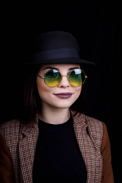 Красивая девушка в шляпе и круглых очках позирует в студии на черном фоне — стоковое фото