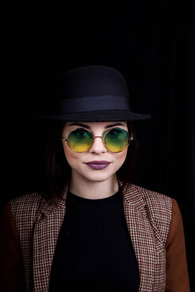 Bella ragazza in cappello e bicchieri rotondi è in posa in Studio su sfondo nero — Foto Stock