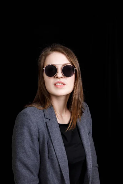 Imagen de una linda joven confiada de pie sobre fondo negro en el estudio, con chaqueta gris y gafas de moda, con el pelo rubio — Foto de Stock