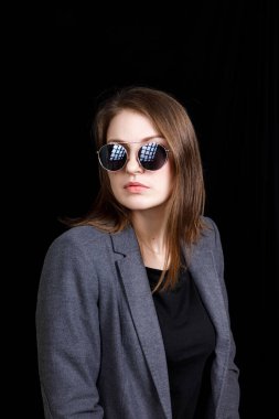 Stüdyoda siyah arka planda duran, gri ceketli, şık gözlüklü, sarı saçlı hoş, kendine güvenen genç bir bayan.