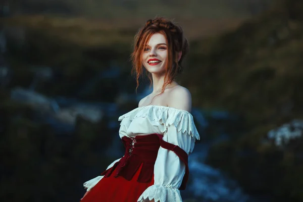 Красивая рыжая девушка в белом и красном костюме улыбается на фоне камней, покрытых мохом и водопадом — стоковое фото