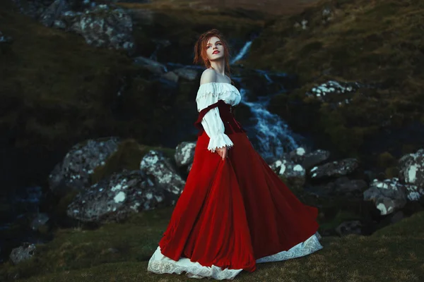 Όμορφη κοκκινομάλλα κορίτσι ντυμένη λευκό και κόκκινο κοστούμι χαμογελά στο παρασκήνιο των λίθων που καλύπτονται με βρύα και καταρράκτη — Φωτογραφία Αρχείου