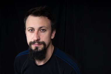 Koyu renk elbiseli, siyah arka planda sakallı genç bir adamın yakın plan portresi.
