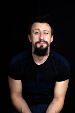 Koyu renk elbiseli, siyah arka planda sakallı genç bir adamın yakın plan portresi.