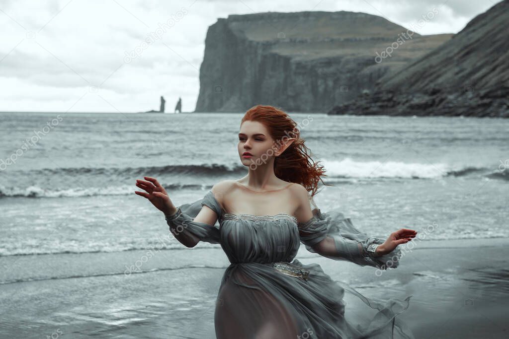 beautiful young girl with elf ears standing on the black sand in the wind near the ocean in a translucent grey dress