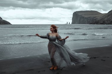 Elf kulaklı güzel bir genç kız okyanusun yakınındaki rüzgarda siyah kumların üzerinde şeffaf gri bir elbiseyle duruyor.