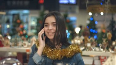 Bir süpermarkette bir neşeli kız portresi. Ağır çekim. Şirin bir kız telefonda konuşuyor. Noel için dekore edilmiş bir süpermarkette bir kız.