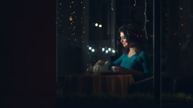 Sevimli kadın bir kafede. Ağır çekim. Güzel bir kadın cam arkasında bir kafede oturuyor ve bir kupa çay dökülen. Kentsel ulaşım ve trafik cam yansıtır.