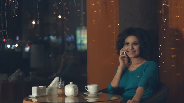 一个咖啡馆里有智能手机的女孩一个漂亮的女孩坐在咖啡馆里 在电话里聊天 她笑着喝着茶 窗外灯火通明的傍晚城 — 图库视频影像