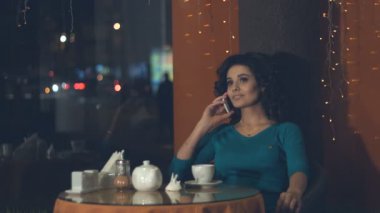 Bir kafede bir akıllı telefon olan kız. Ağır çekim. Güzel bir kız bir kafede oturan ve Evet. O gülümsüyor. Dış pencere akşam şehrin ışıkları.