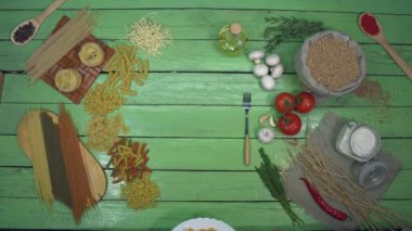 Tabakta ıspanaklı makarna. Üst Manzara. Yeşil masa sol tarafta birçok çeşit kuru makarna ve sebzeler, kiraz domatesleri, otlar, un ve sağ tarafta mantarlar olmak üzere ikiye ayrılır..