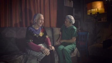 Yaşlılara iyi bak. 2 Shot.1. Bir sağlık çalışanı akşamleyin bir odada yaşlı bir kadınla konuşuyor. Şaka yapıyorlar ve gülümsüyorlar. Yakın plan. Hemşire büyükannesinin elini okşar..