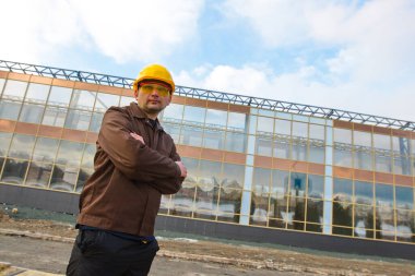 Mühendis ya da inşaatçının portresi. Tulumlu, koruyucu kasklı ve gözlüklü bir adam. Arkasında modern cam bir bina var..