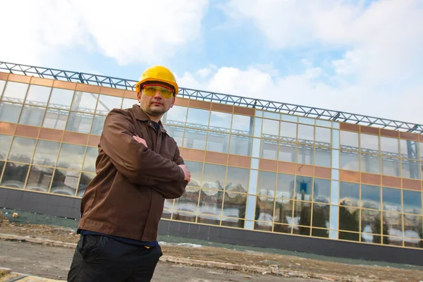 Mühendis ya da inşaatçının portresi. Tulumlu, koruyucu kasklı ve gözlüklü bir adam. Arkasında modern cam bir bina var..