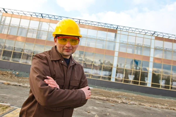Portrait of an engineer or builder. A man (worker, engineer) in overalls, in a protective helmet and glasses. Behind him is a modern glass building.