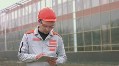 Mühendis ya da inşaatçının portresi. Önlüklü ve koruyucu başlıklı genç bir adam (işçi, mühendis) tablet kullanır. Arkasında modern cam bir bina var. Kar yağıyor.