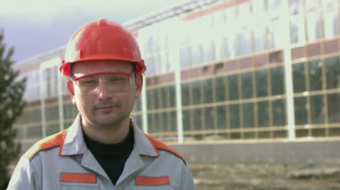 Mühendis ya da inşaatçının portresi. Ağır çekim 2x. Koruyucu miğfer ve gözlük takan bir adam. Arkasında modern cam bir bina var. Kamera sağdan sola hareket ediyor..