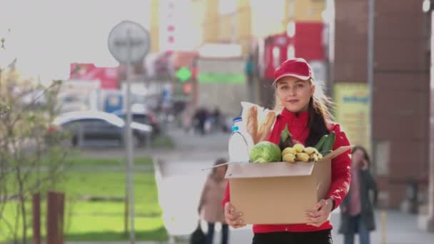 一个带着一盒食物在街上的女孩 送货服务的女孩拿着一个盒子 盒子里有蔬菜 她把盒子举到前面笑了 — 图库视频影像