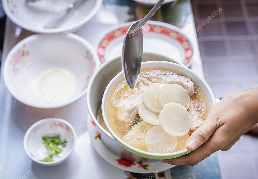 Close up of white radishes on pork bone, yellow wonton soup in top view with a hand holding a green bowl and blurred background, Asian homemade style food concept.