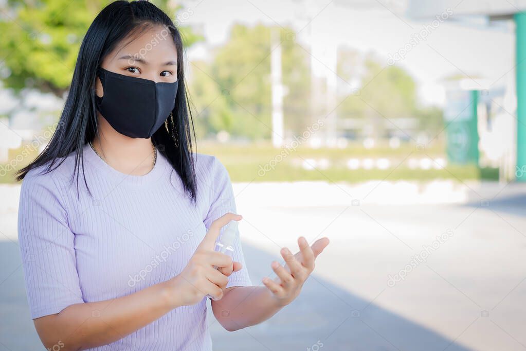 A cute young Asian woman wears a black mask and uses alcohol spay to clean a hand for preventing the epidemic of Covid-19. There is a copy space on the right, the concept of healthcare, and hygiene.