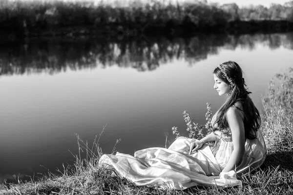 Красивая девушка в длинном платье сидит на берегу реки и смотрит вперед. Черно-белое фото — стоковое фото