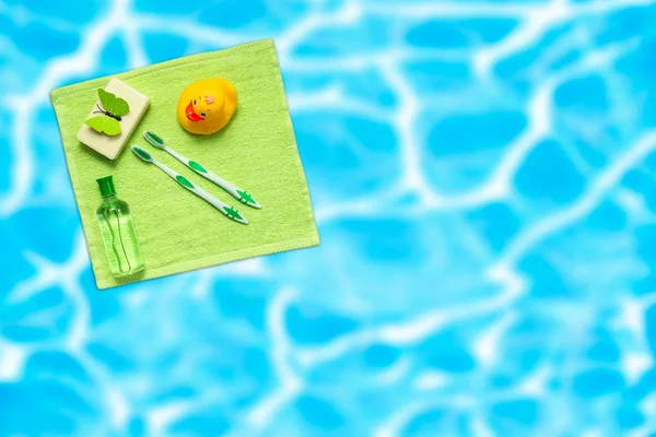 Acessórios de banheiro em uma toalha verde. Pato de borracha, escovas de dentes, sabão e loção. Fundo de água da piscina. Higiene das crianças . — Fotografia de Stock