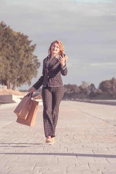 Joyeux beau blond souriant femme avec bretelles courir avec des sacs en papier et téléphone à la main le long de la promenade — Photo