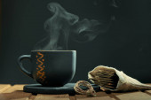 Horizontální záběr šálek horkého nápoje s párou a sáček čajových sáčků na dřevěném stole nad černým pozadím.