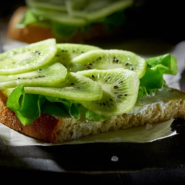 Sandwich con queso suave y kiwi en rodajas de cerca en la tabla de cortar sobre fondo oscuro. Concepto de desayuno saludable . — Foto de Stock