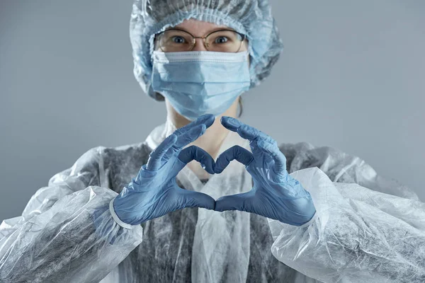 Horizontal shot of young woman in glasses with a medical mask and hands in latex glove shows the symbol of the heart.