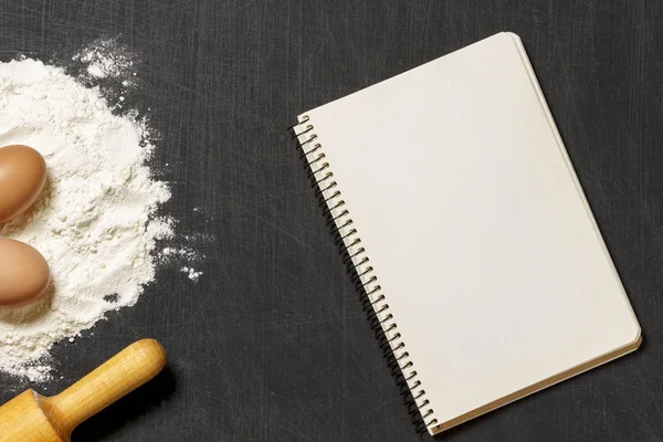 Hamur için malzemeler. Un, yumurta, oklava ve siyah scratchet arka plan üzerinde boş not defteri. Üstten Görünüm. Düz yatıyordu. Mockup menü veya tarifi.