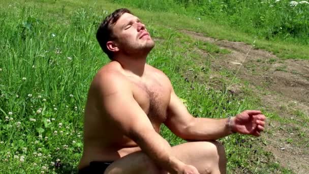 Man relaxed and tans with eyes closed sitting on a green meadow on a sunny day — Stock Video
