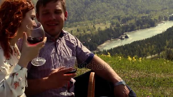 Pár élvezi a szabadban, bort iszik, ül egy zöld rét közepén magas hegyekben — Stock videók