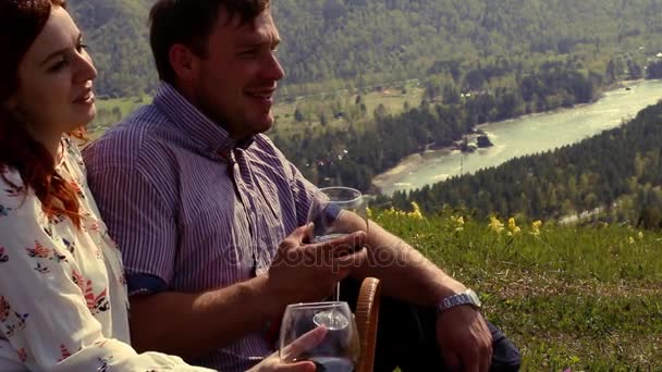 Pareja disfrutando del aire libre, bebiendo vino, sentada en un prado verde en lo alto de las montañas — Vídeos de Stock