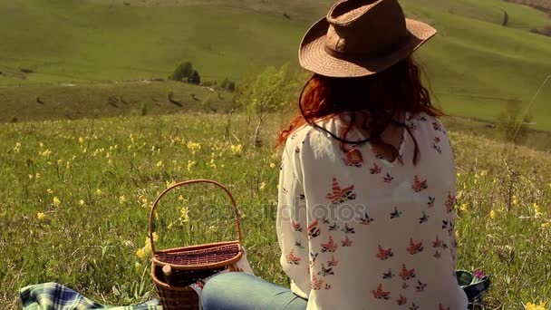 Mujer en un sombrero bebiendo vino sentada en un prado verde en lo alto de las montañas — Vídeo de stock