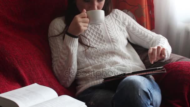 Молода жінка п'є каву і читає електронну книгу, сидячи біля вікна — стокове відео