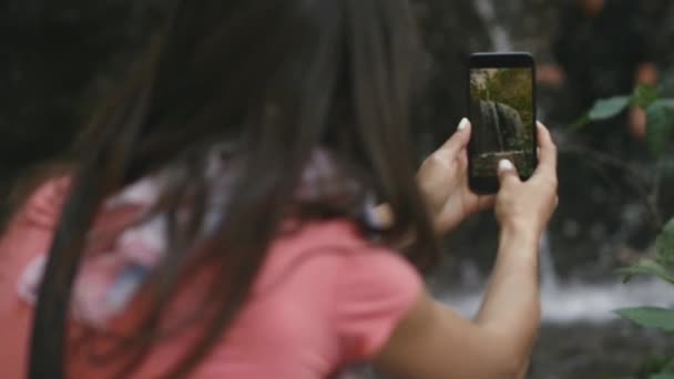 Mujer joven toma una cascada en un teléfono inteligente mientras viaja — Vídeos de Stock