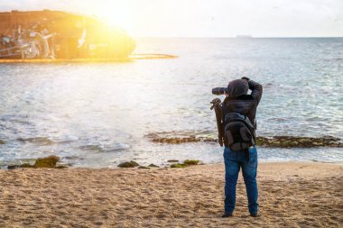 Baharda kumsalda fotoğraf makinesi ve sırt çantasıyla gezen gezgin, hala soğuktayken fotoğraf çeker.