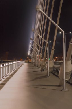 Kaldırım ve otoyol, büyük çelik kablolarla dolu bir köprüden geçiyor. Gece vakti parlak ışıklarla kapanıyor.