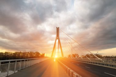 Büyük çelik kablolarla kablo destekli bir köprüden geçen otoyol, akşamları yakın plan gökyüzü, bulutlar ve güneş ışınlarıyla çevrili.