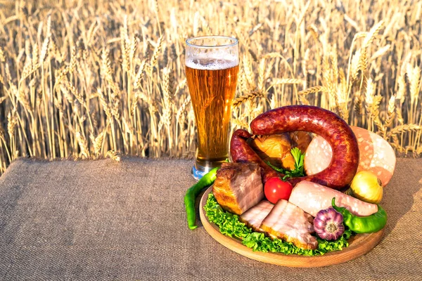 Beer, meat products. Smoked ham, sausage, bacon, cheese, vegetables lying on a table in a wheat field against the blue sky